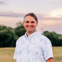 Head-and-shoulders outdoor portrait of Kasey Zientek, HSE Product Analyst at MCG Energy, standing in a grassy field, wearing a light-colored short-sleeve button-down shirt with a blue floral pattern. The person is facing the camera. The background is softly blurred, showing green trees and an evening or early morning sky with pale pink, purple, and cream tones. Natural lighting creates a bright, professional-looking photograph.